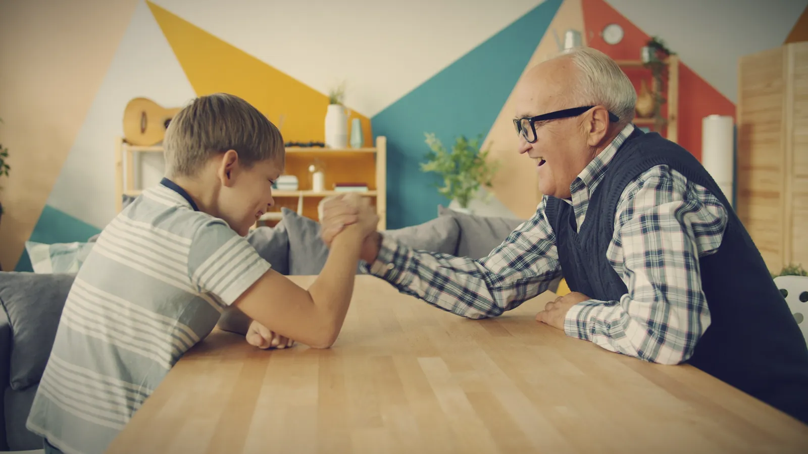 A boy in a striped T-shirt arm-wrestles a smiling grandfather across a wooden table in a brightly painted living room