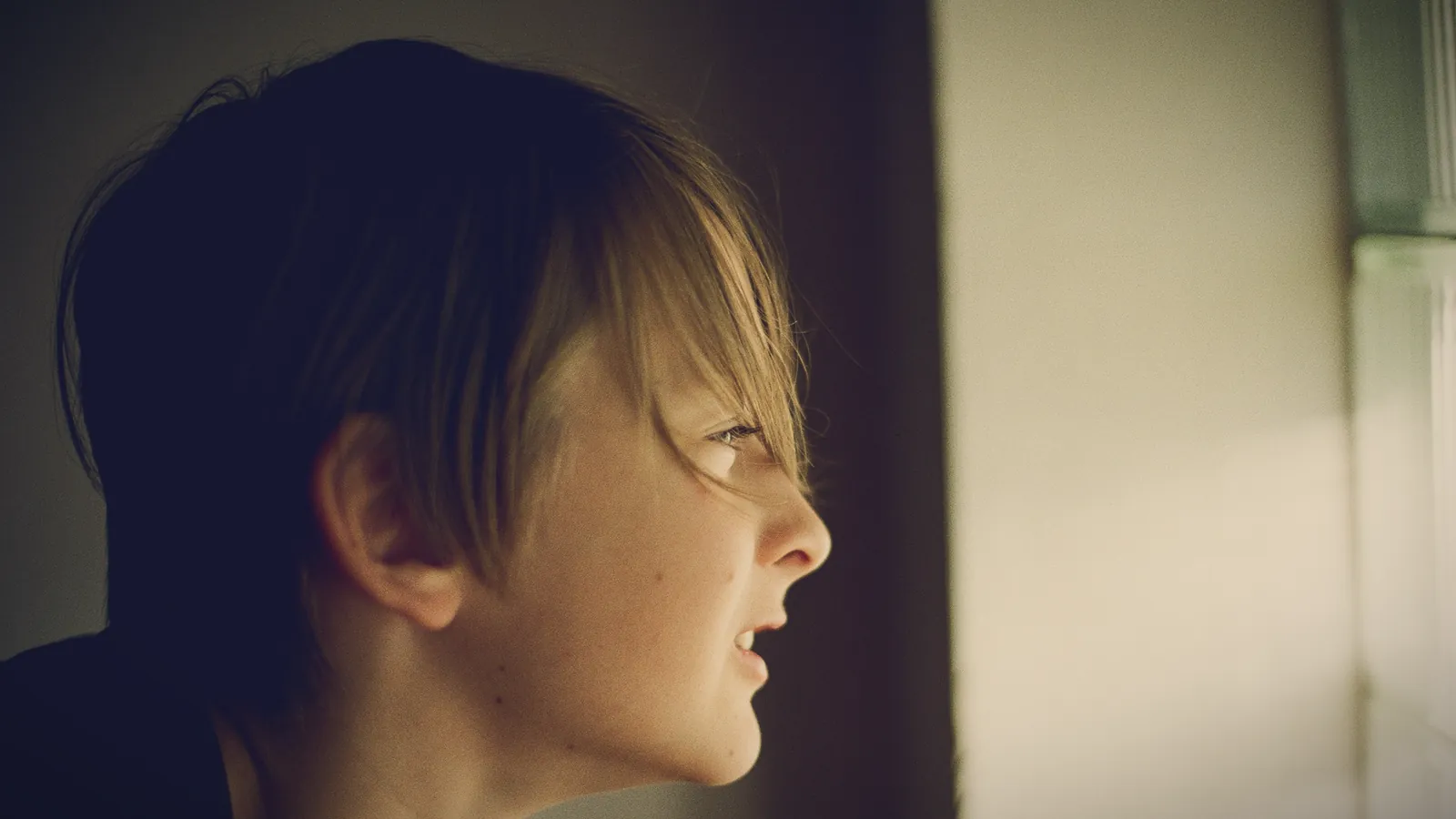 A child with a shaggy fringe, seen in profile, wrinkles their nose while looking toward a bright window