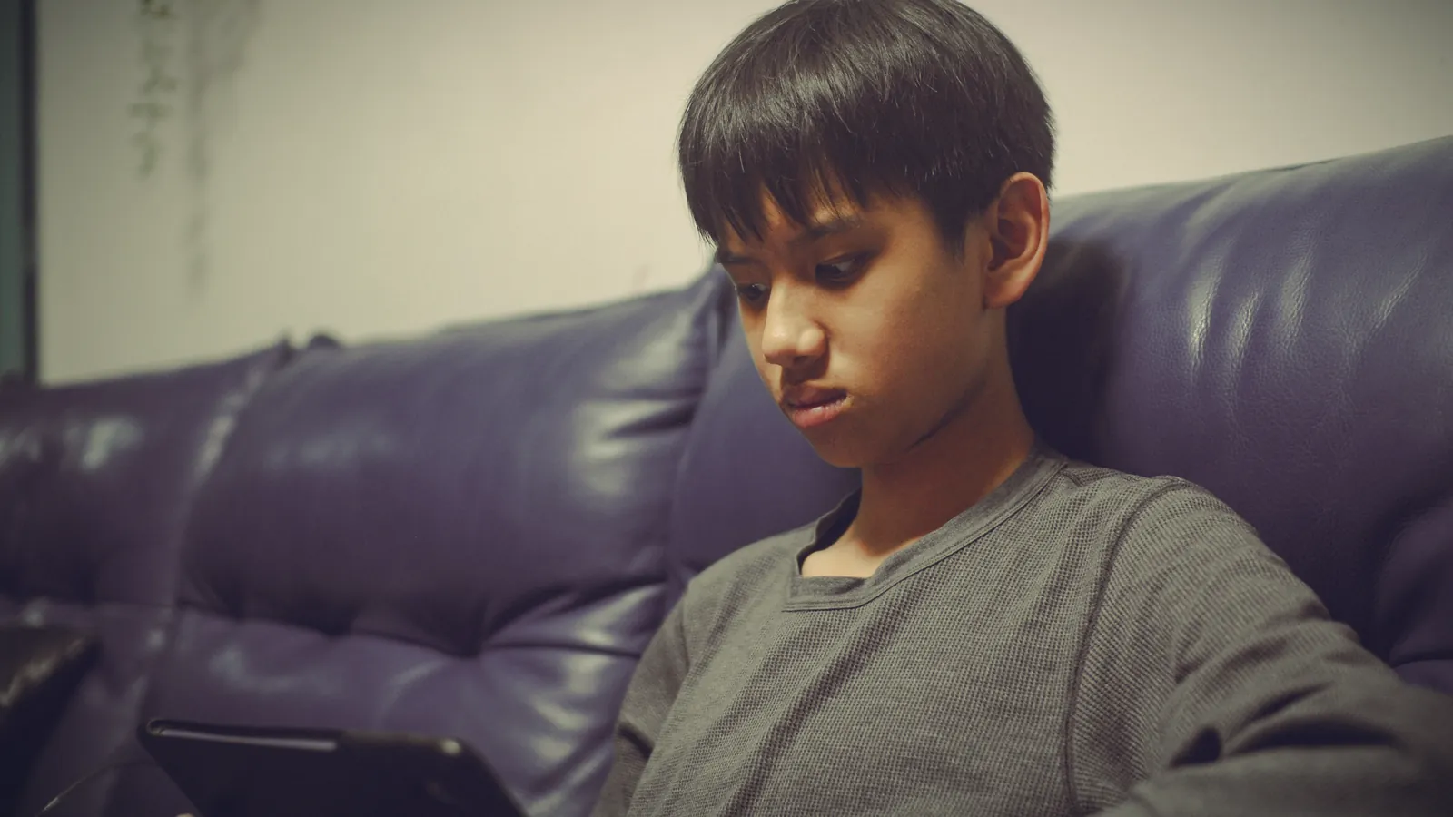 A preteen boy slumped on a leather sofa with a tablet, looking sullen
