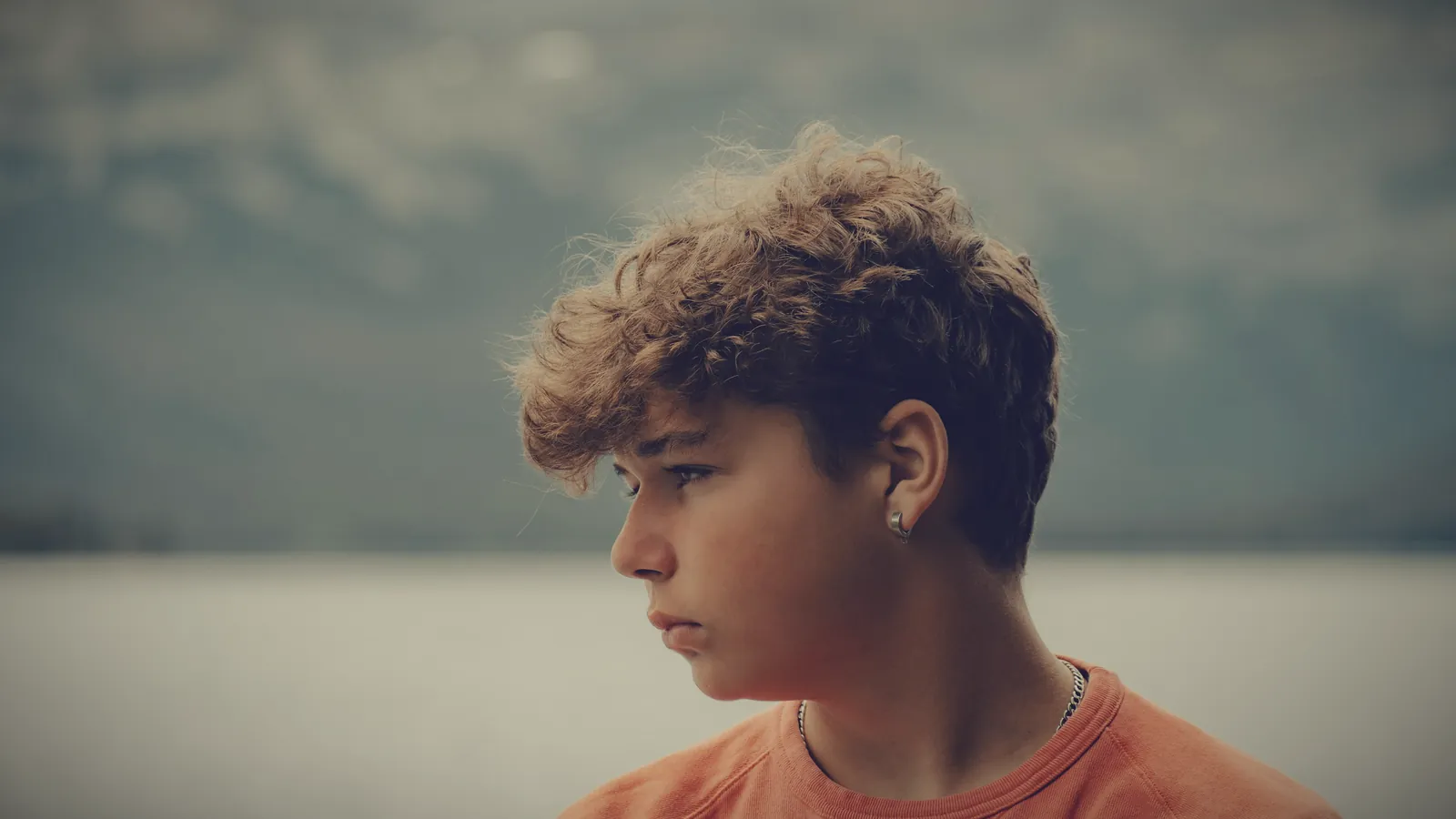 A teenage boy with curly hair looking away, jaw set, by a lake