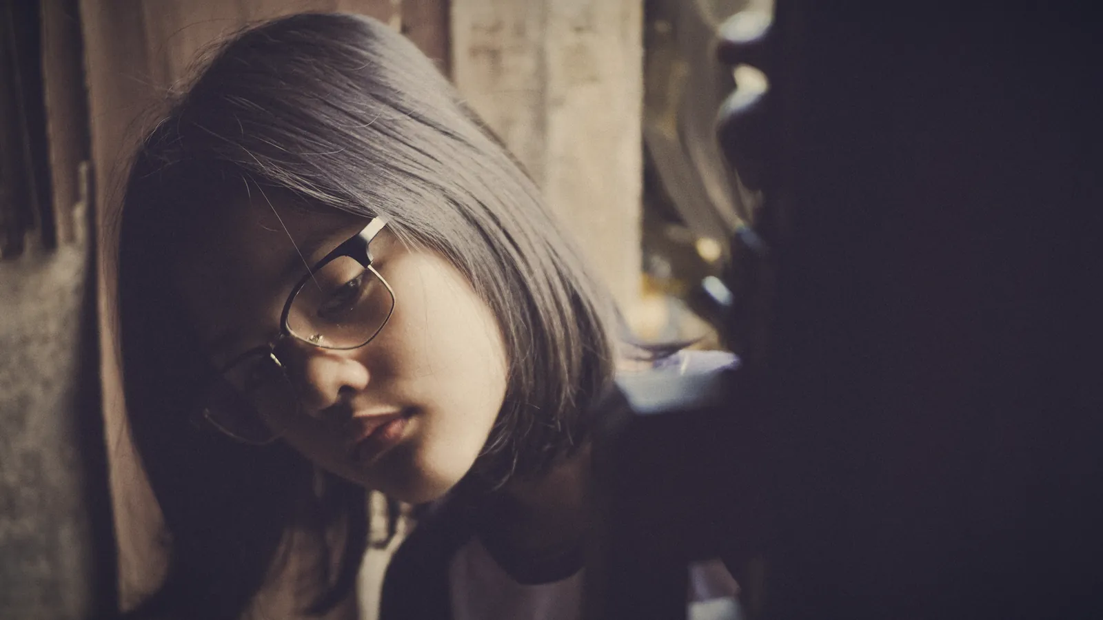 A teenage girl with glasses and shoulder-length dark hair leans forward and looks down, half in shadow