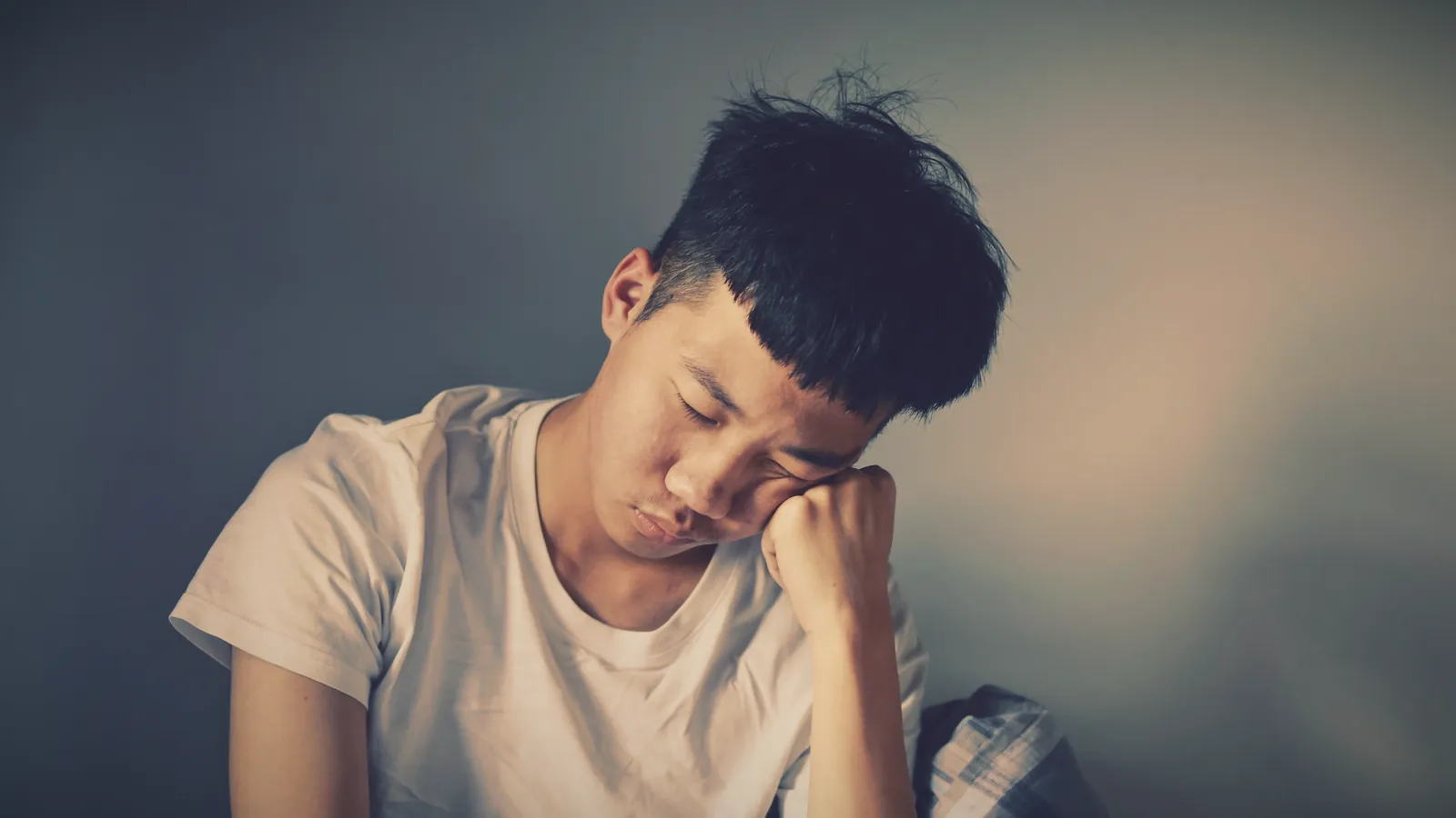 A teenage boy in a white T-shirt sits with his eyes closed, resting his cheek on his fist against a grey wall