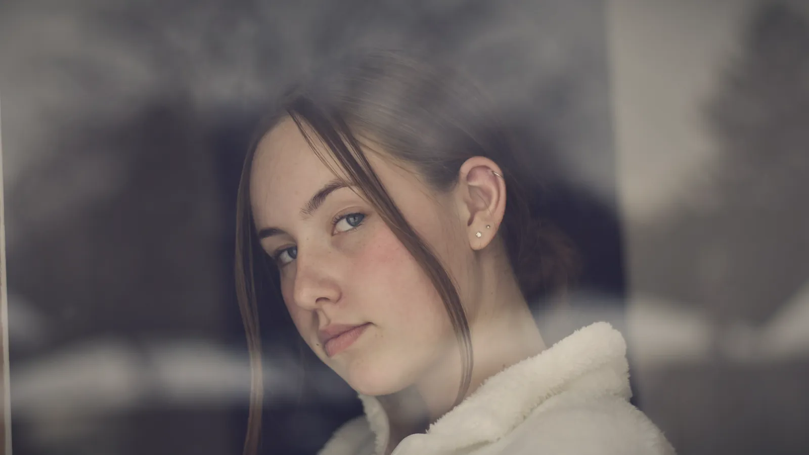 A teenage girl in a white fleece jacket looks out through a window pane at the camera