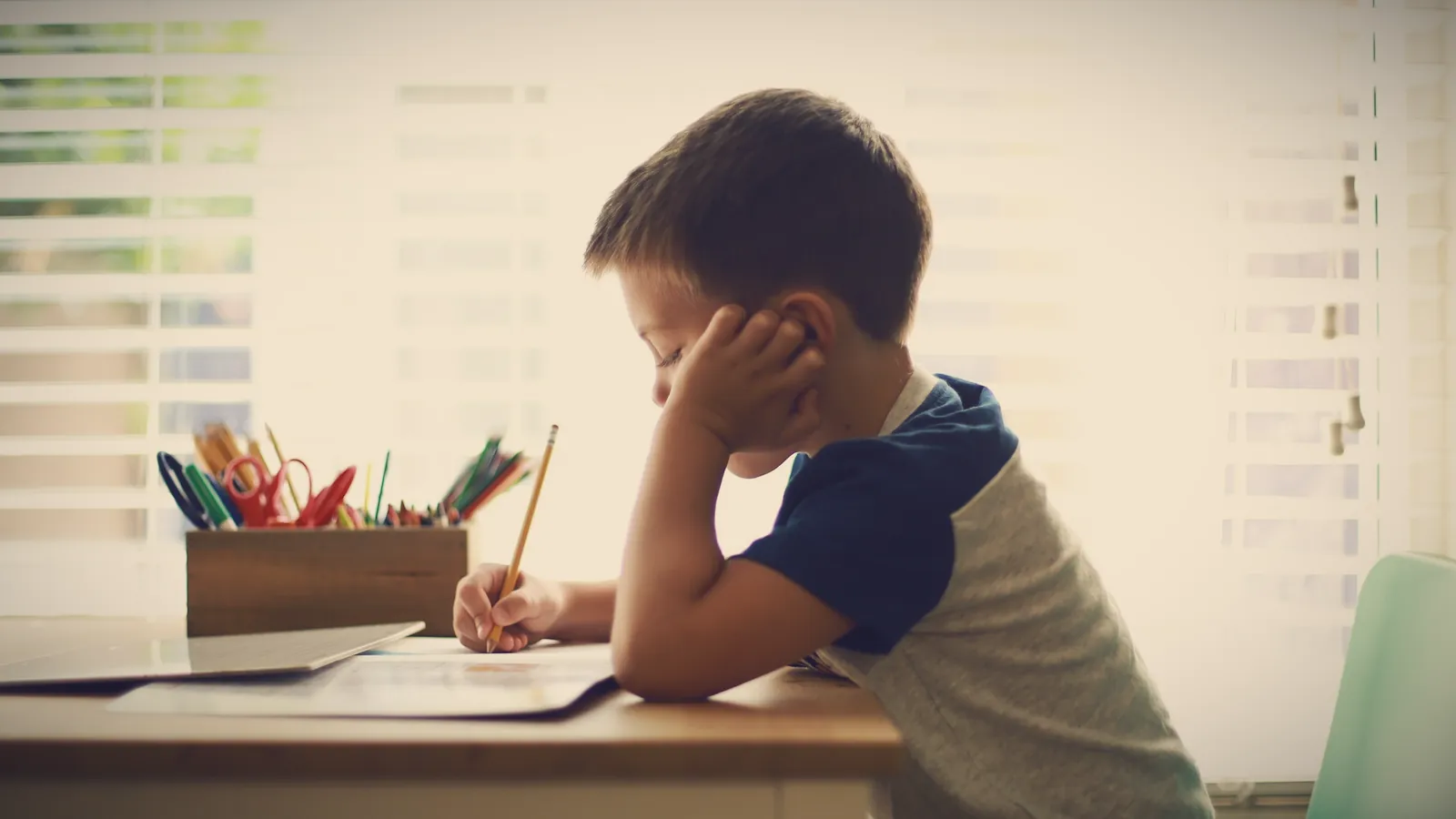 A young boy at a desk by the window, head in his hand, struggling through his homework