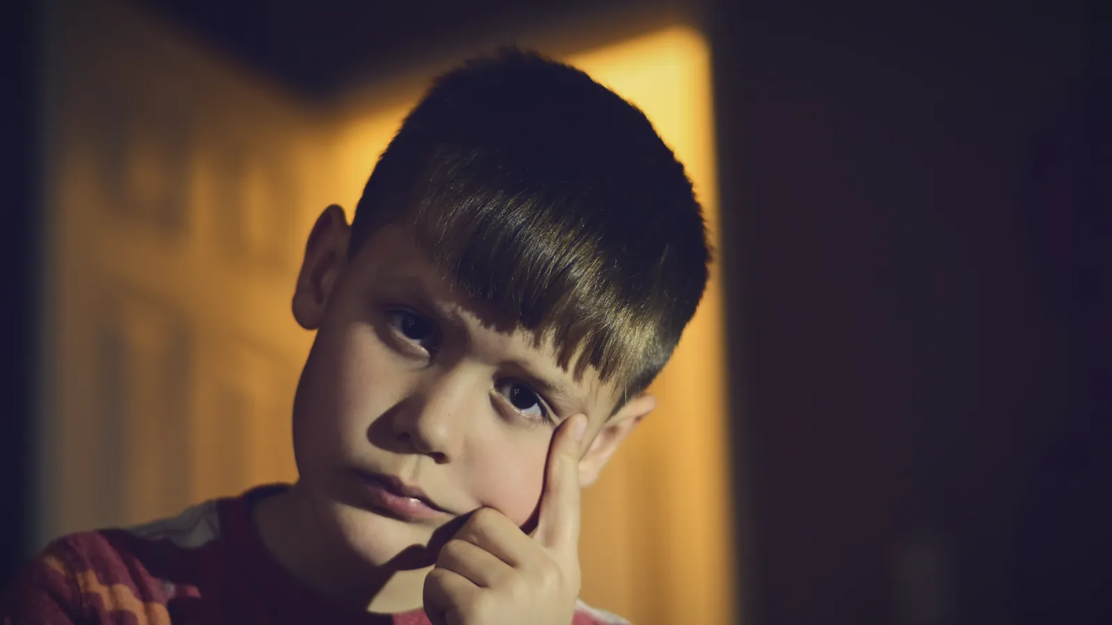 A boy with short brown hair frowns slightly and rests a finger against his cheek in warm indoor light
