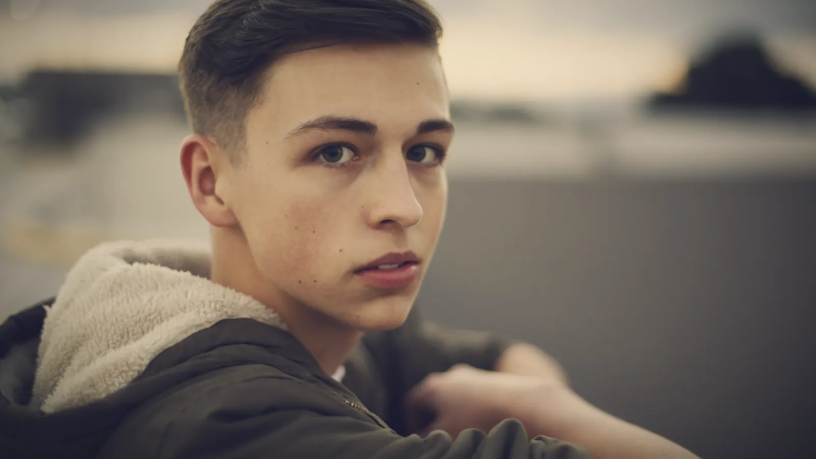 A teenage boy in a hooded jacket looking back over his shoulder with a hard, guarded stare