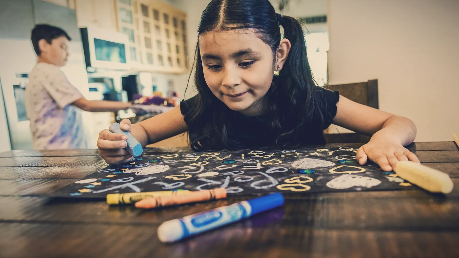A girl drawing with colorful markers on a sheet at the kitchen table
