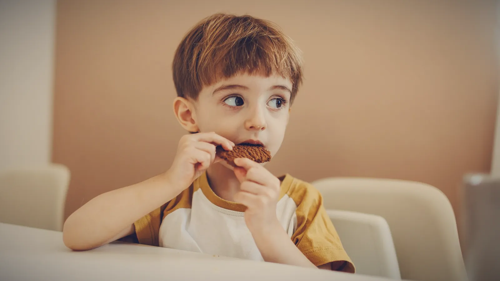 A little boy at the kitchen table biting a cookie and glancing sideways, hoping nobody noticed