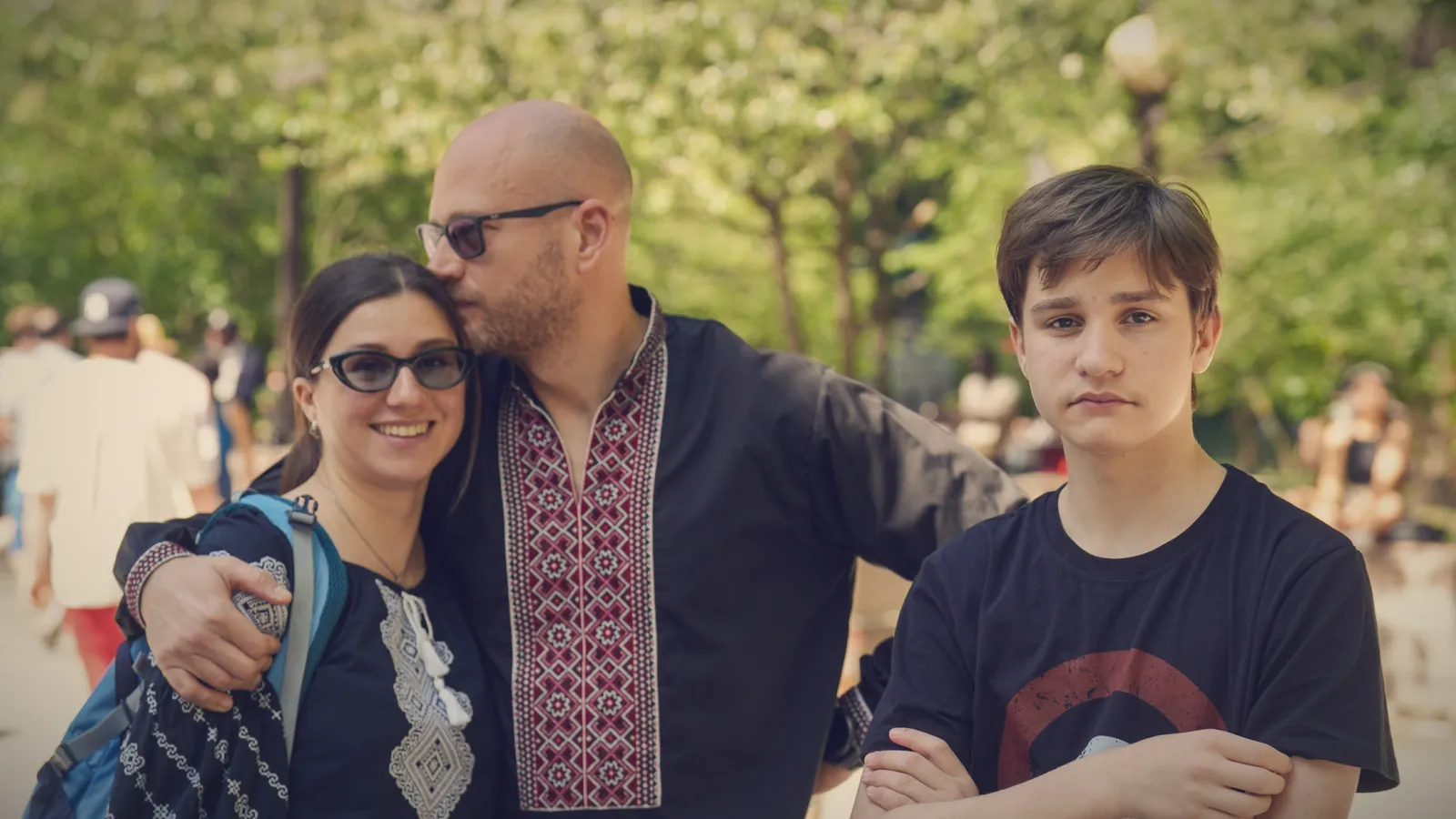A teenage boy standing with his arms crossed and a sullen look, a little apart from his parents