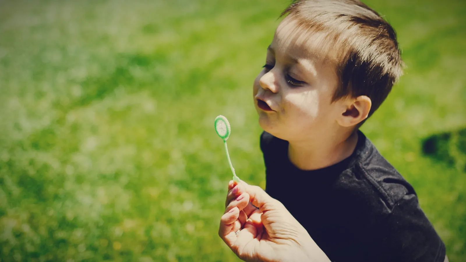 A young boy taking a slow breath to blow a bubble from a wand his parent is holding