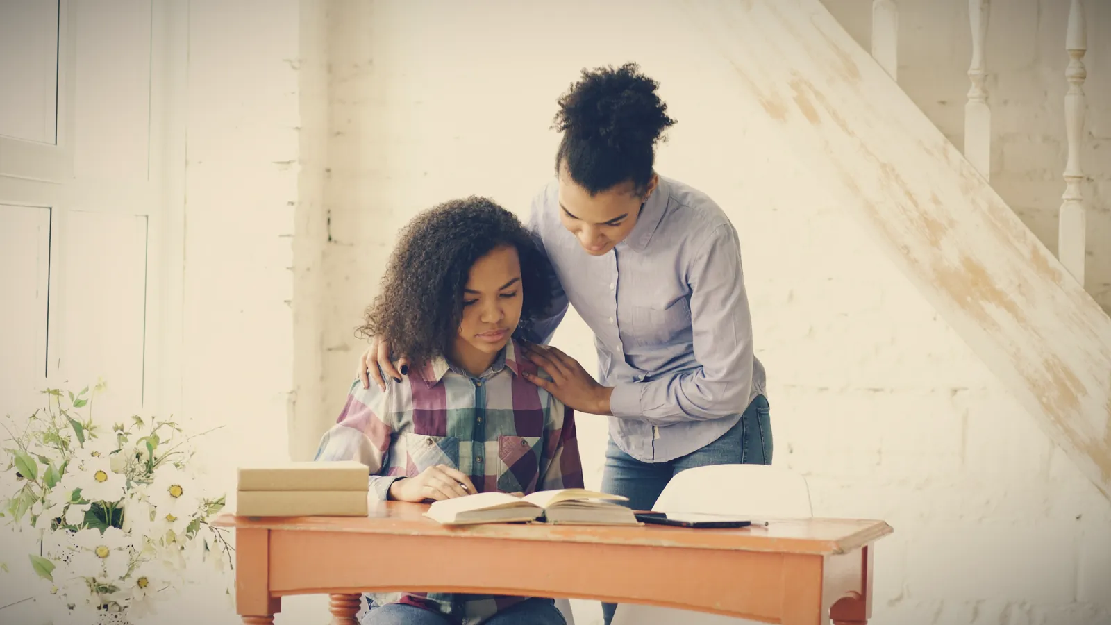 A woman standing beside a teenage girl at her desk, one hand resting on her shoulder