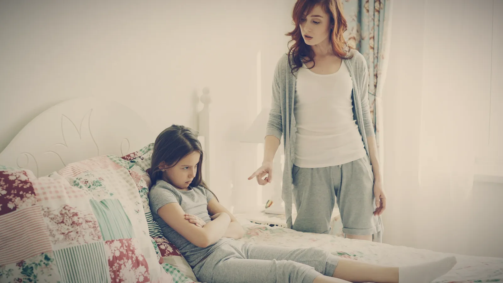 A mother pointing and scolding while her daughter sulks on the bed with her arms crossed