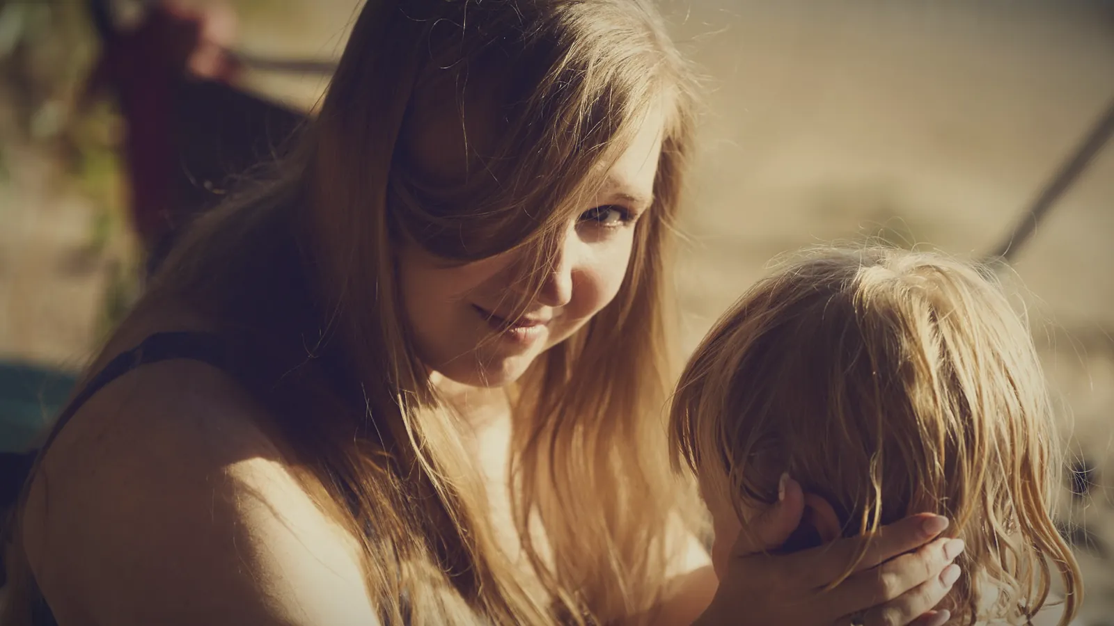 A mother smiling gently at her young daughter, who is hiding her face in her hands
