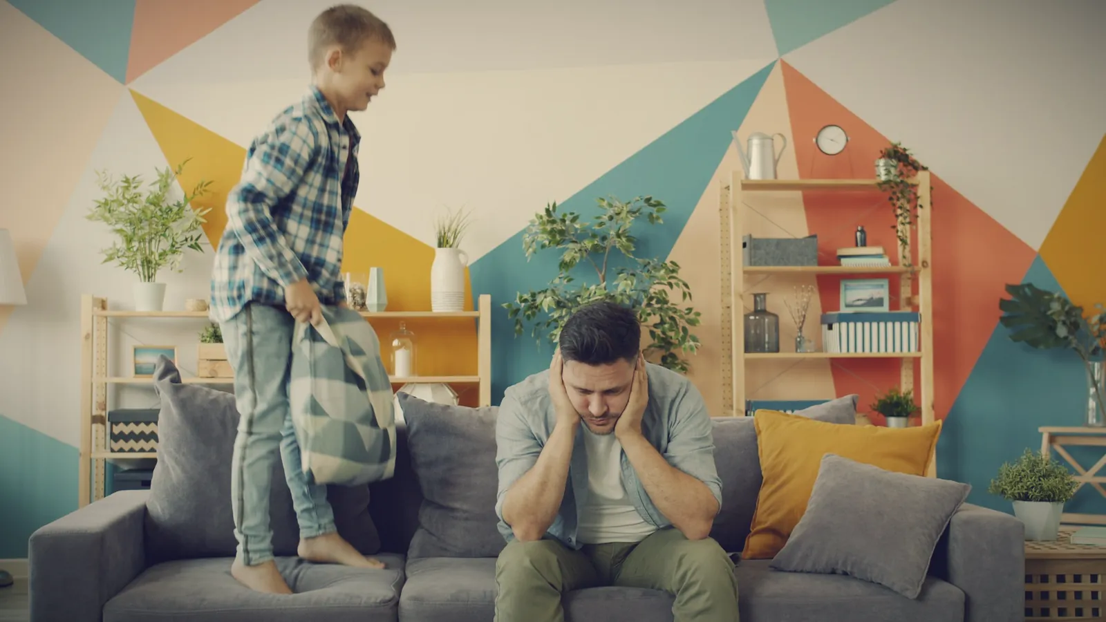 A worn-out father holding his head on the sofa while his son jumps beside him with a pillow
