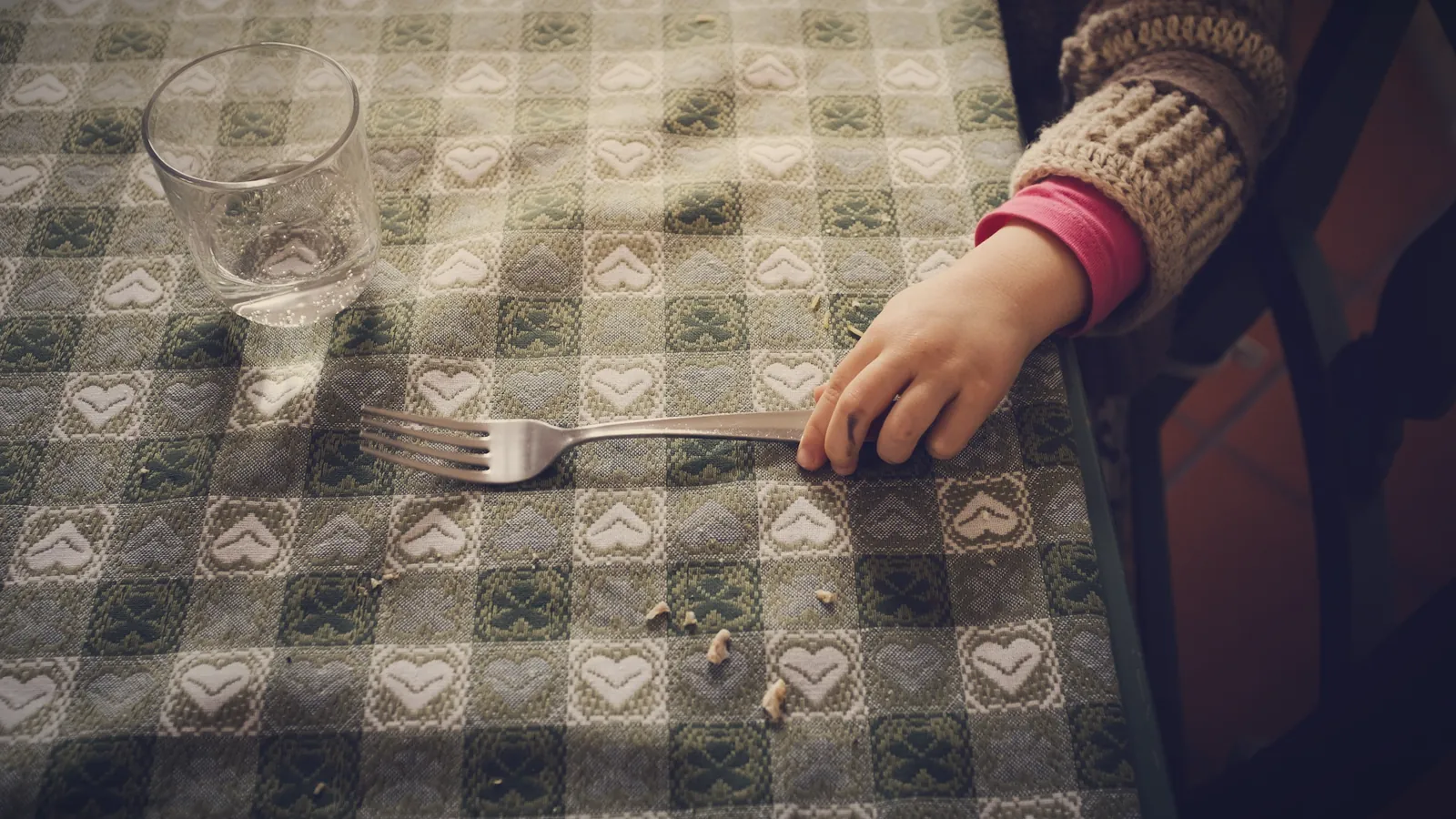 A child's hand resting next to a fork, a glass and a few crumbs on a checked tablecloth