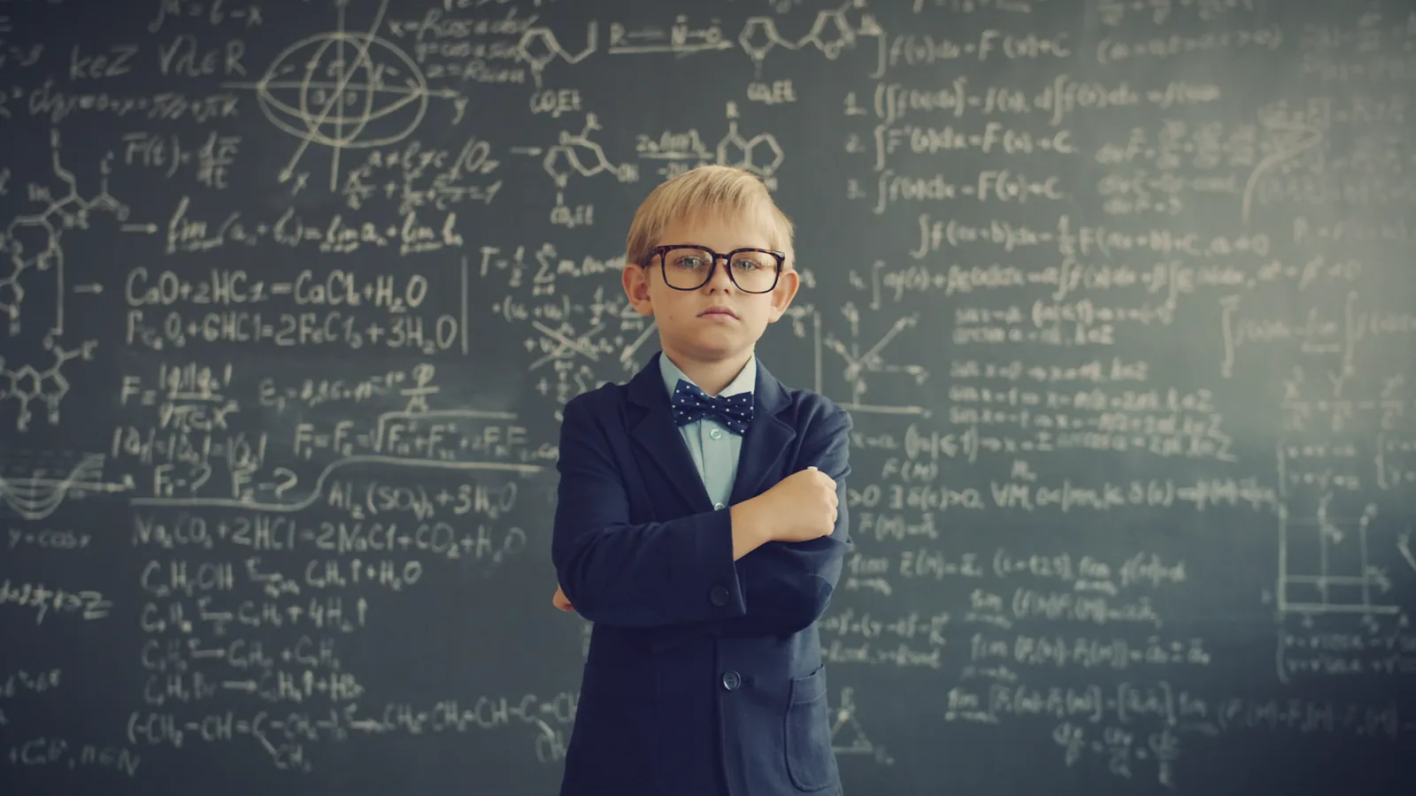 A confident little boy in a suit and glasses standing with his arms crossed in front of a chalkboard