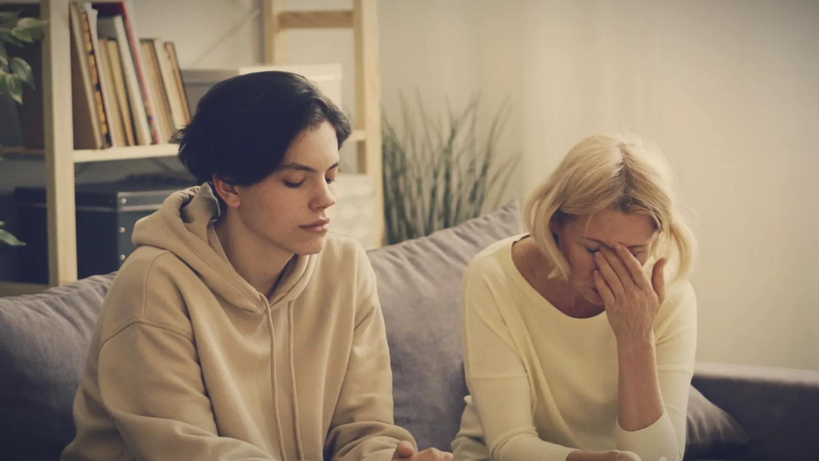 A teenage girl sitting stiffly on the sofa, eyes down, beside her mother who is in tears