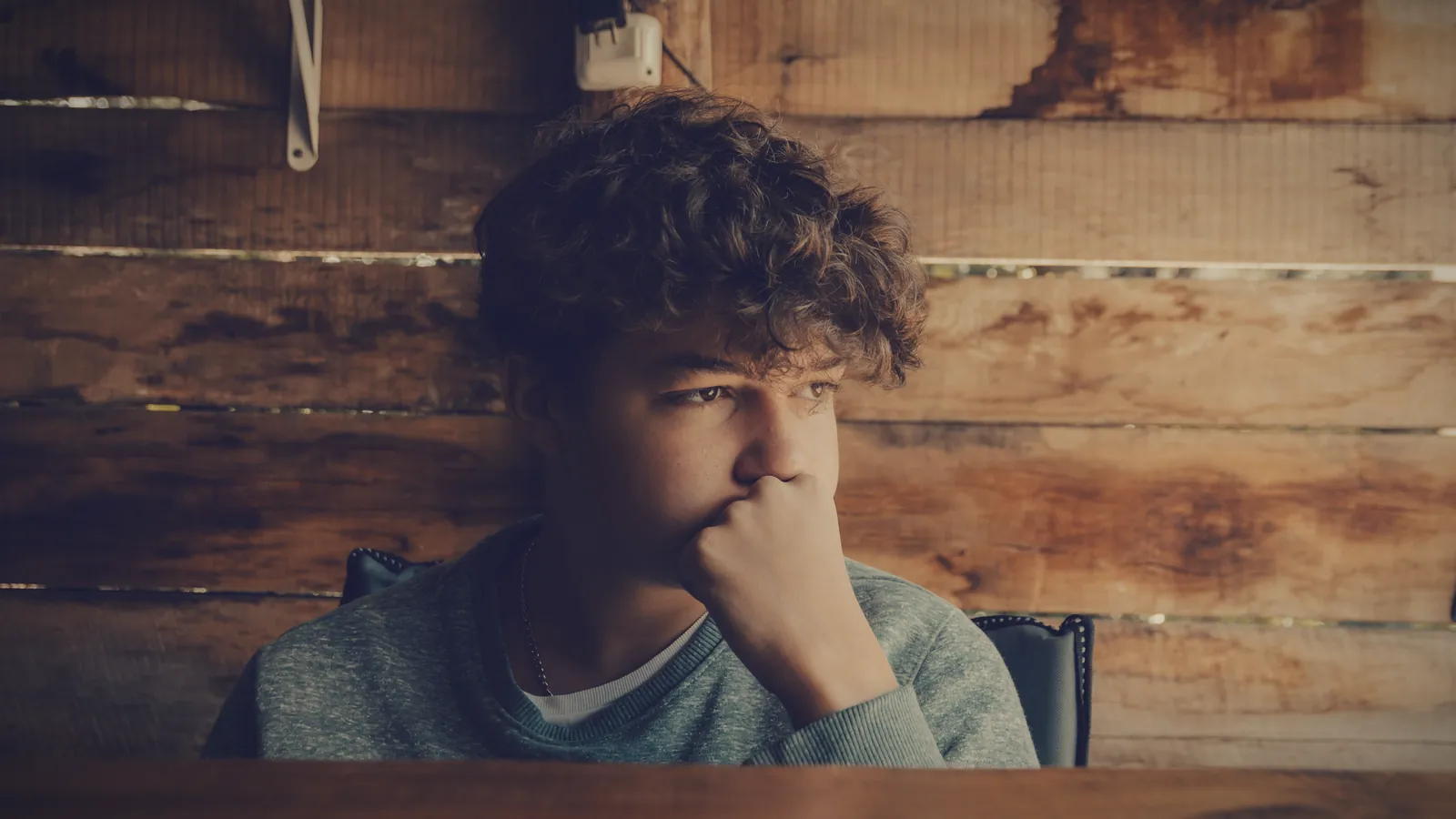 A teenage boy at a wooden table, hand over his mouth, looking away