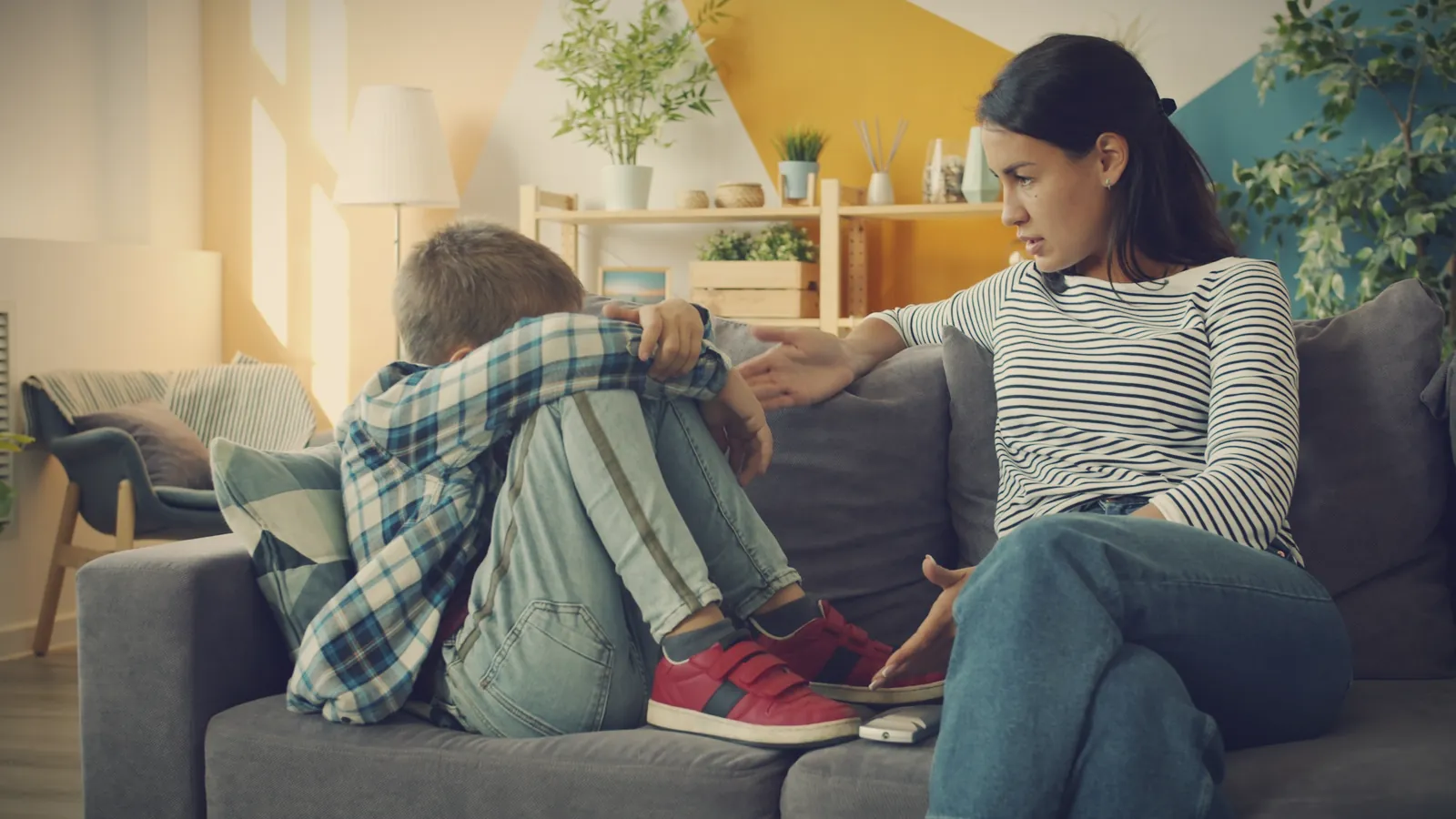 A boy curled up on the sofa at home while his mother sits beside him and reaches out
